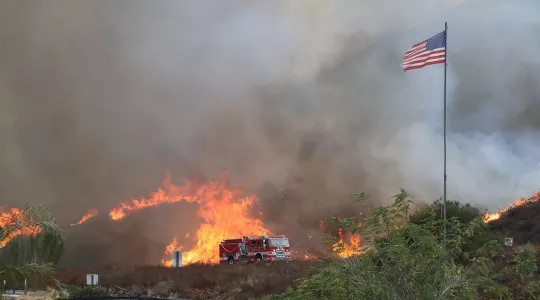 Image of a fire apparatus driving in front of a hillside engulfed in flame.