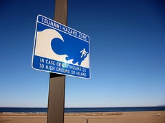 Shows a signpost with the image of a wave, stating:TSUNAMI HAZARD AREA. in case or earthquake go to higher grounf